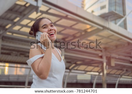 Smartphone woman pretty asian in white dress talking on phone at afternoon in city around Skyscraper with sunset. Beautiful young female professional having casual conversation on mobile phone. 