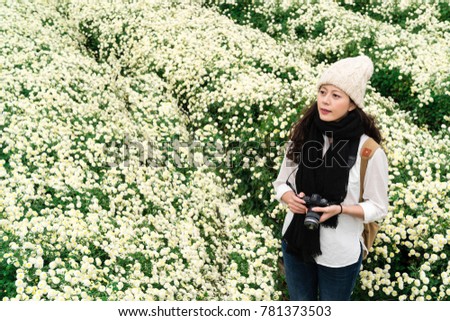 Portrait of beautiful asian woman with wool cap and scarf holding camera in the white chrysanthemum garden in autumn. Stand and gaze at the scenery.