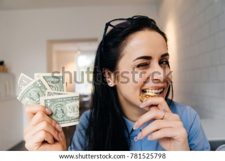 funny brunette girl in blue shirt tries gold bitcoin to taste, holds dollars in her hand