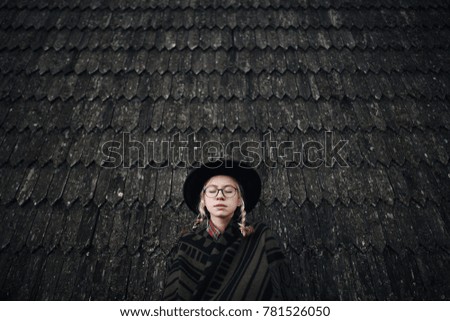 Young blonde girl in a hat, poncho and glasses near the wooden house. Autumn picture. Lifestyle. Background