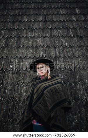 Young blonde girl in a hat, poncho and glasses near the wooden house. Autumn picture. Lifestyle. Background