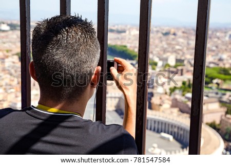 man takes a picture of St. Peter's Square in the Vatican on his smartphone through a lattice on the dome of St. Peter's Cathedral 