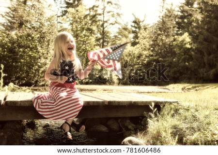 Portrait of happy little Caucasian girl wearing dress with stars and stripes sitting on wooden bridge waving American flag at and laughing at Independence day