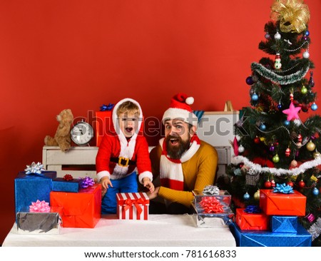 Christmas eve concept. Santa and little assistant among gift boxes near fir tree. Christmas family opens presents on red background. Man with beard and surprised face plays with boy on New Year eve