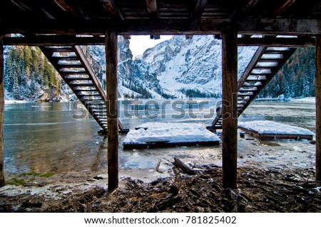 small wooden staircase framed by the poles that support the refuge towards the mountain lake in the snow with blue skies and snow-capped mountains
