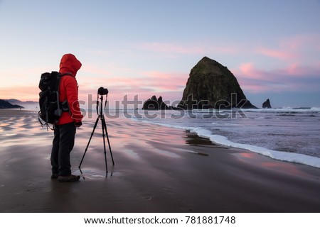 Photographer with a camera is standing on the sandy beach during a vibrant and colorful winter sunrise. Taken in Canon Beach, Oregon Coast, United States of America. Royalty-Free Stock Photo #781881748