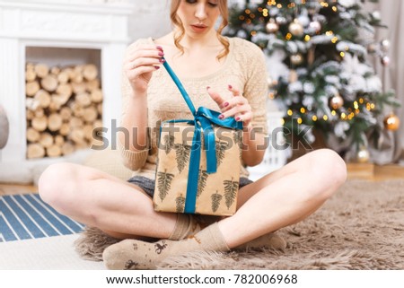 the girl sits next to a tree and a fireplace and opens Christmas presents