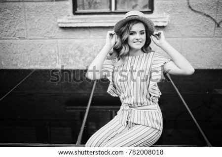 Portrait of a fabulous young woman wearing striped overall and hat sitting on black shiny surface next to the building. Black and white photo.