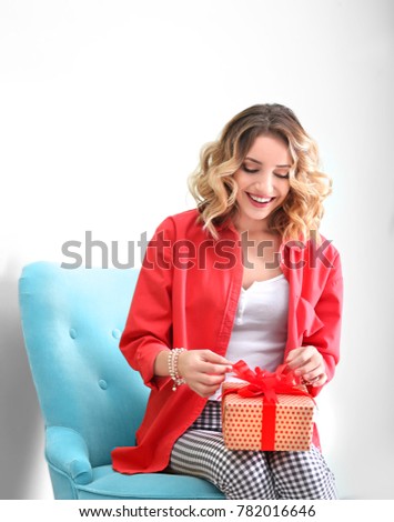 Attractive young woman with gift box sitting in armchair near color wall
