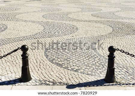 A view of a wavy cobblestone plaza with black security chains and poles in the foreground, Lisbon, Portugal