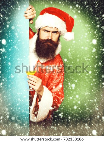 handsome bearded santa claus man with long beard on serious face holding glass of alcoholic beverage in christmas or xmas sweater and new year hat on colorful studio background