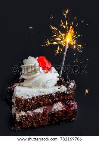 A holiday birthday or Christmas background image - a piece of Black Forest cake with a bright red candy on top and a burning Bengal fire on a dark gray background