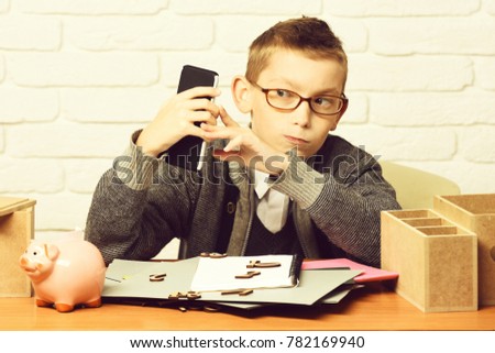 young cute businessboy in grey sweater and glasses sitting at table with papers pink piggy pig bank and holding cell phone on white brick wall background