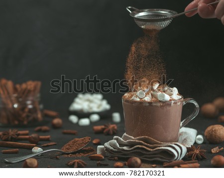 Hand sprinkled cinnamon powder on glass mug with hot chocolate cocoa drink. Copy space. Dark background. Low key. Winter food and drink concept.