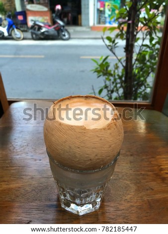 Hot coffee with heart shape latte art milk foam on top in coconut shell  over clear glass on wooden table top with street view