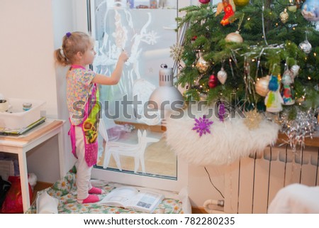Little girl draws snow pictures on the window with stencil and white paint