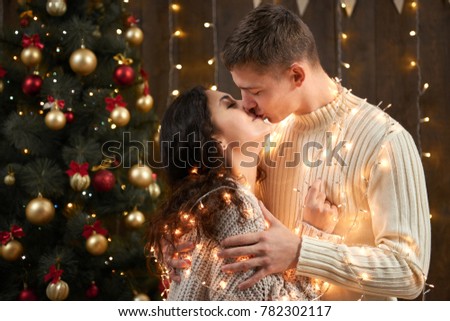 young couple kissing in christmas lights and decoration, dressed in white, fir tree on dark wooden background, romantic evening, winter holiday concept