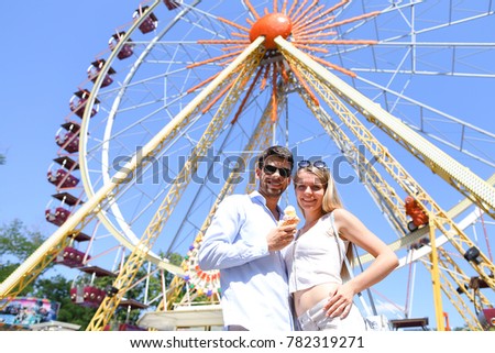 Cute young people, newlyweds kissing, hugging and smiling, looking into eyes of each other and posing for photos, guy holding ice cream on background of large and colorful ferris wheel at an amusement