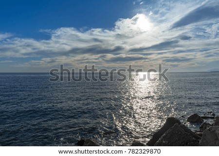 A blue sky with the sun behind the clear clouds above the ocean.