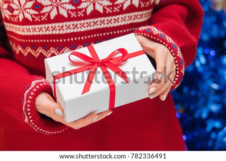Woman showing beautiful white gift box with red ribbon- closeup of present on holiday sweater background and Christmas tree.