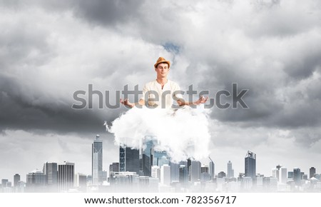Man in white clothing keeping eyes closed and looking concentrated while meditating on cloud above wooden floor with cityscape view on background.