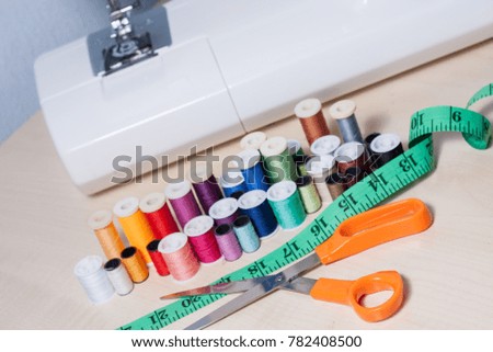 Colourful collection of home sewing accessories: tailor shears or scissors, flexible green measuring tape, multiple asorted sewing threads or yarn in colors of the rainbow, next to a sewing machine.