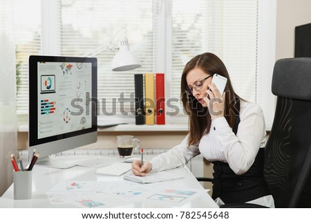 Beautiful business woman in suit and glasses sitting at the desk with cup of coffee, working at computer with documents in light office, talking on mobile phone resolving issues, looking aside