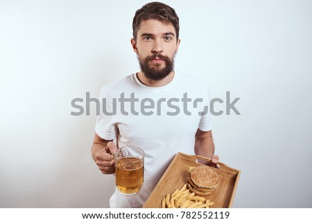 man with a beard holds a beer and a tray with fast food on a light background                              