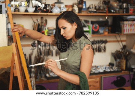 Girl-artist paints a painting on canvas. Canvas stands on the easel. In the background, shelves with accessories for drawing