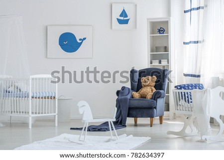 White baby room interior with a rocking horse on white rug and a teddy bear on navy blue armchair