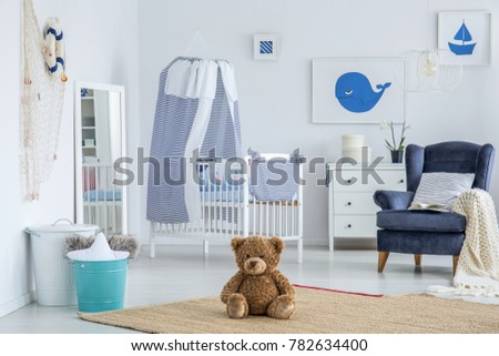 Striped cushion and beige blanket lying on a navy blue armchair next to white wooden crib in bedroom for a child