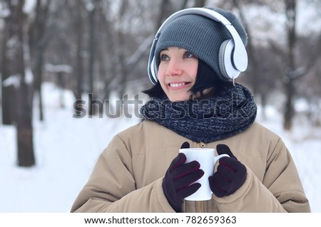 Winter portrait of pretty attractive caucasian pierced girl in snowy wood. She is listening to music on big white headphones with coffee cup in her hands. Walks in the open air