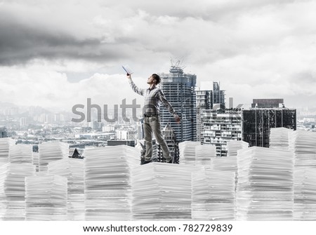 Young man in casual wear keeping hand with book up while standing on pile of documents with cityscape on background. Mixed media.