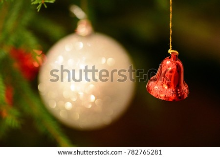 Christmas tree with small red hanging bell decoration and white ball decoration in background. Blurred soft focused background. Warm lighted picture.  Focus on ball decoration