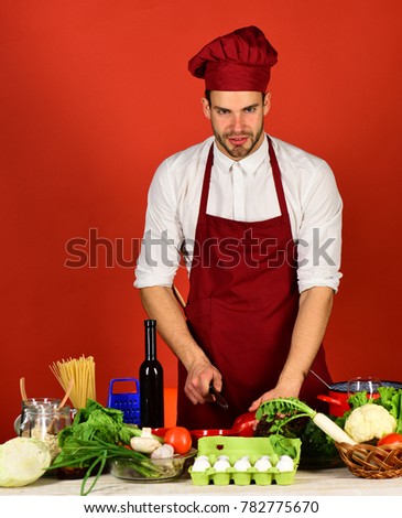 Chef with smiling face holds knife and red bell pepper on red background. Man in cook hat and apron chops pepper. Food preparation concept. Cook works in kitchen near table with vegetables and tools.