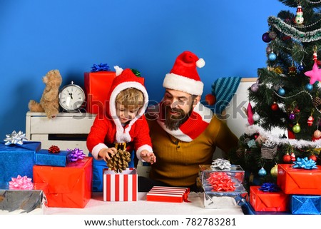 Santa and little assistant among gift boxes near Christmas tree. Boxing day and family concept. Man with beard and cheerful face plays with son. Christmas family opens presents on blue background