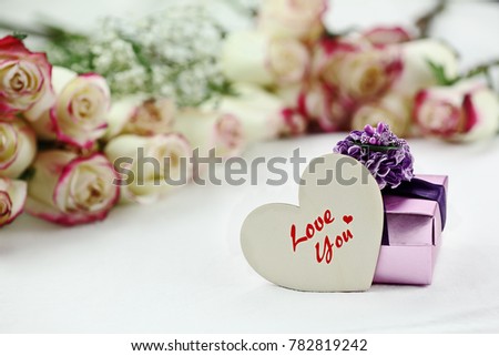 Gift box and Love You wooden heart card in front of a beautiful bouquet of  red and white roses in the background. Shallow depth of field with selective focus on heart.