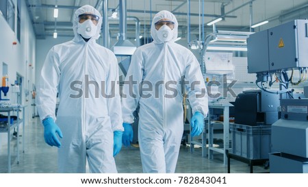 Two Engineers/ Scientists in Hazmat Sterile Suits Walking Through Technologically Advanced Factory/ Laboratory. Clean High-Tech Environment with CNC Machinery. Royalty-Free Stock Photo #782843041