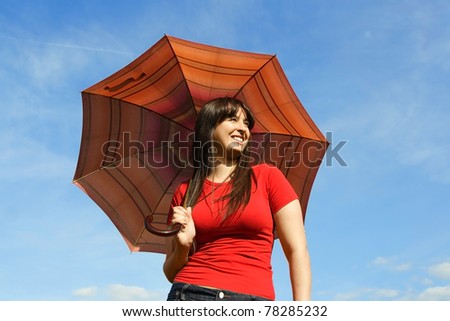 Young brunette girl in red shirt holding red umbrella and smiling, blue sky