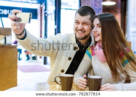 Happy young couple making a selfie at the restaurant. Drinking tea or coffee, smiling and taking a photo with mobile phone. Winter dating of man and woman.