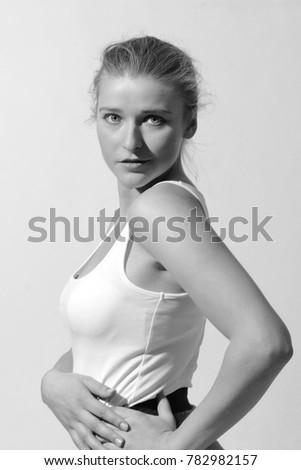 A beautiful girl is posing in a photo studio with a white background. She is wearing a white tank top and grey sports skirt. With exposed shoulders. She has nice brown hair in a bun. 