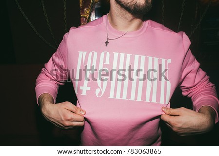 A young man with a beard dressed in a pink sweatshirt with the inscription "Sinner". On the neck hangs a chain with the Orthodox cross. Conceptual photography