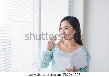 Beautiful asian girl drinking water and reading book near window at home