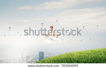 Young man in white clothing keeping eyes closed and looking concentrated while meditating on cloud in the air with city view on background.