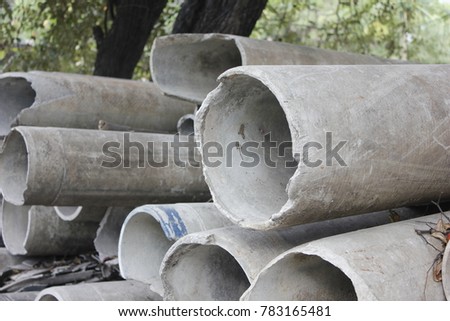 Stack of grey cement tubes on gravel ground with tree in background