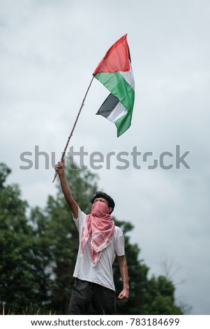 A male teenager covering his face with a keffiyeh or serban while holding a Palestine flag