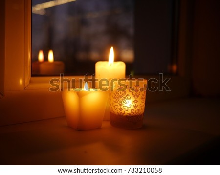 Photo of three lit candles - two paraffin wax candles and one tealight in a glass candle holder with glittery golden ornament, standing on a windowsill late in the evening.