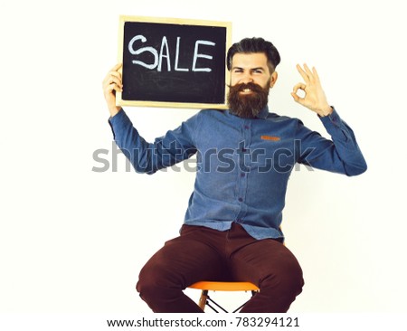 Bearded man, long beard. Brutal caucasian amused hipster with moustache holding sale inscription on blackboard, sitting on orange chair wearing blue shirt and vinous pants isolated on white background
