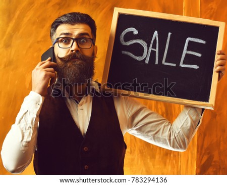 Bearded man, long beard. Brutal caucasian unshaven surprised hipster with glasses and moustache talking by cell phone, holding sale inscription on blackboard on brown studio background
