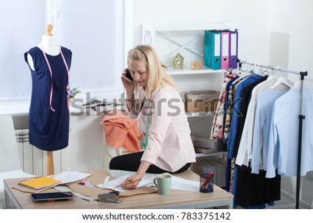 Communication with suppliers by phone, skype. Beautiful European woman tailor talking on the phone, standing near the table in the workshop with clothes hanging in the background.
